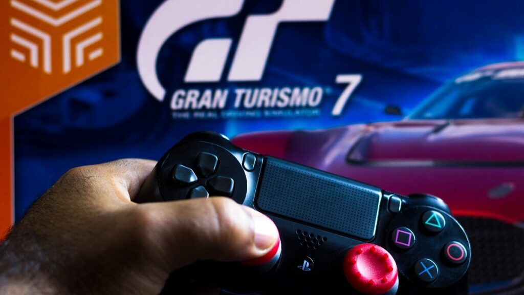 Lista dos melhores jogos de corrida para PS4, foto de uma mão segurando um controle preto e vermelho na frente de um pôster de Gran Turismo 7